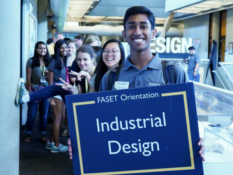 A FASET advisor giving new students a building tour and holding up a sign with the words "Industrial Design".